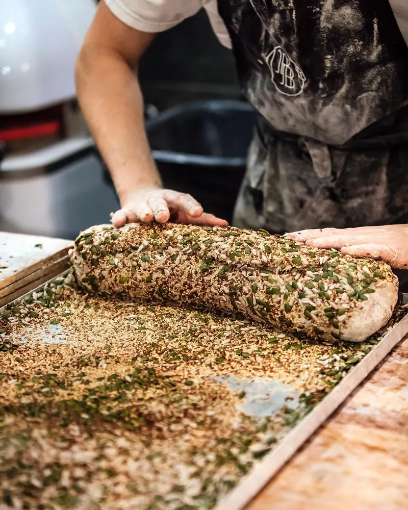 A seeded loaf being rolled through a tray of grains